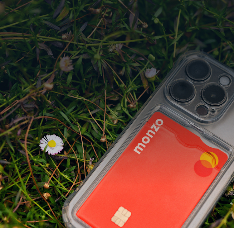 A Monzo card is visible through a clear phone case, which is lying on a grassy surface with daisies nearby.