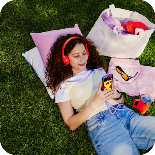 A woman lies in a blanket with headphones on, looking at her phone