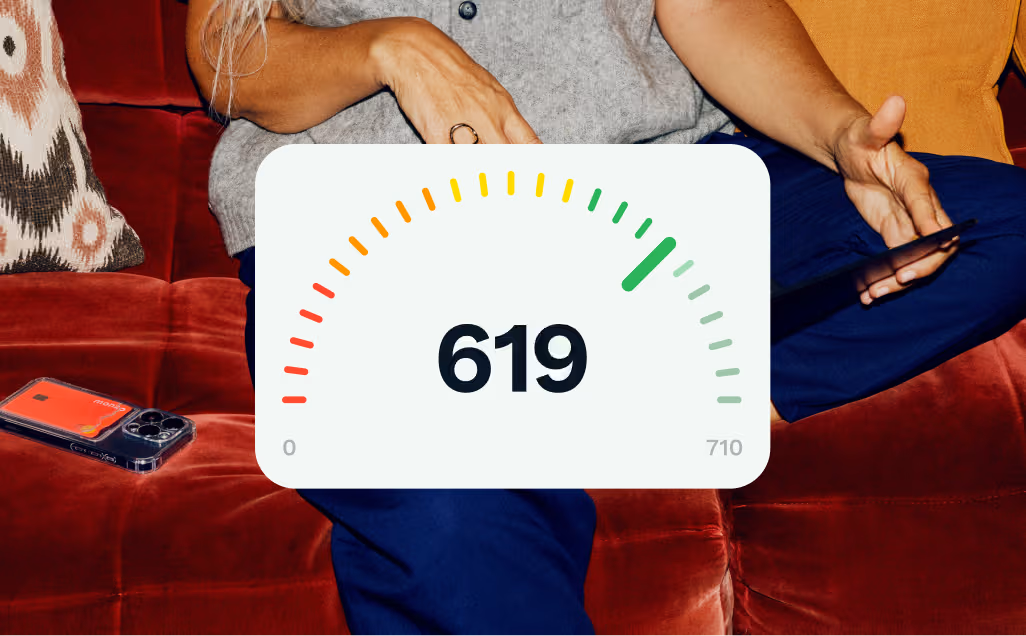 A person sits on a burgundy velvet couch looking at a laptop. Overlaid is a large white semi-circular gauge displaying the number 619 in bold. The gauge’s colour-coded scale transitions from red on the left, through orange and yellow, to green on the right, with the needle pointing to the green zone near the upper right. The scale ranges from 0 on the left to 710 on the right.