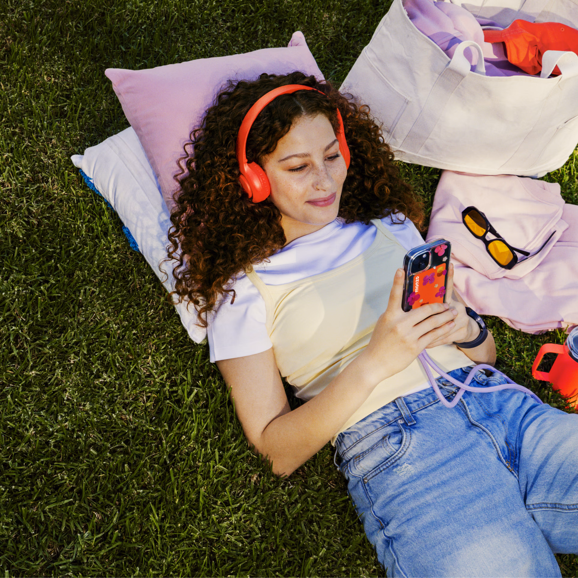A teenage wearing hot coral headphones lies on the grass, looking at a smartphone with a hot coral Monzo card visible in the phone case. A tote bag and sunglasses rest nearby.