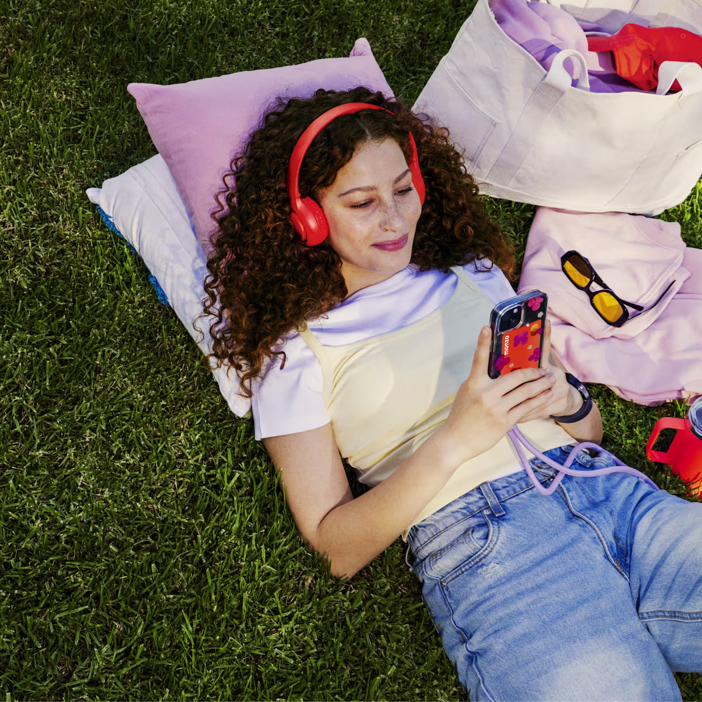 A teenage wearing hot coral headphones lies on the grass, looking at a smartphone with a hot coral Monzo card visible in the phone case. A tote bag and sunglasses rest nearby.