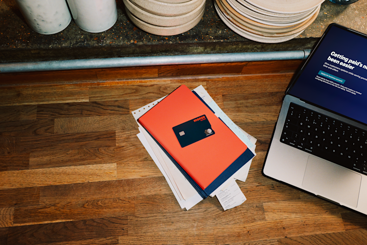 Overhead view of a restaurant workspace with a Monzo business card resting on paperwork beside a laptop, stacked plates visible on the shelf above.