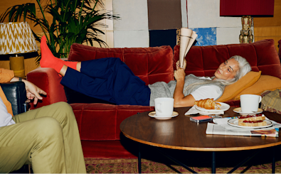A woman reclined on a red sofa and reading a newspaper, talking to someone in an adjacent chair who we can't really see. The coffee table holds pastries, coffee, more newspapers, and a phone with a Monzo card visible in the case