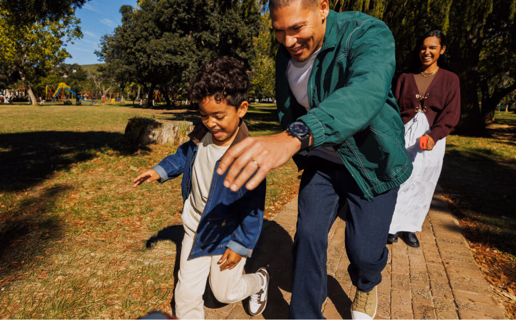 A family with young children enjoying a walk together through a sunlit, grassy park surrounded by trees.