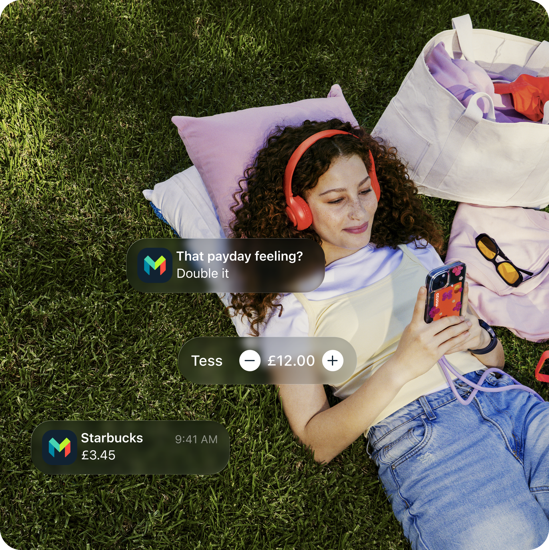 Young girl lying on the grass listening to music in her Hot Coral headphones. She's holding her phone with her Monzo card in the phone case.