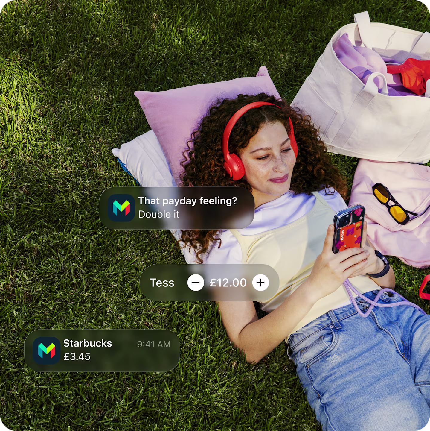 Young girl lying on the grass listening to music in her Hot Coral headphones. She's holding her phone with her Monzo card in the phone case.