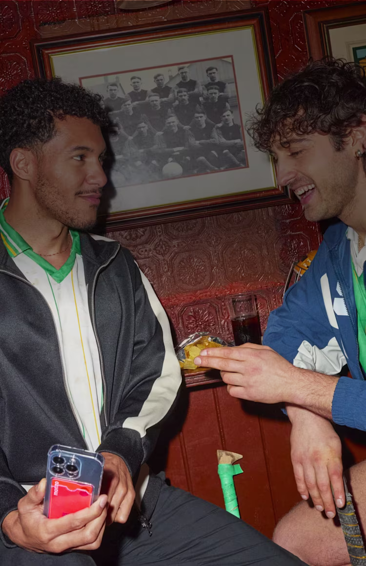 Two friends chatting in a traditional pub with vintage football memorabilia on the walls. One holds a phone while the other holds a drink, sharing a lighthearted moment.