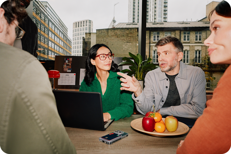 Four business owners having a friendly meeting around a table in a bright office with city views, discussing plans over a laptop and fresh fruit.