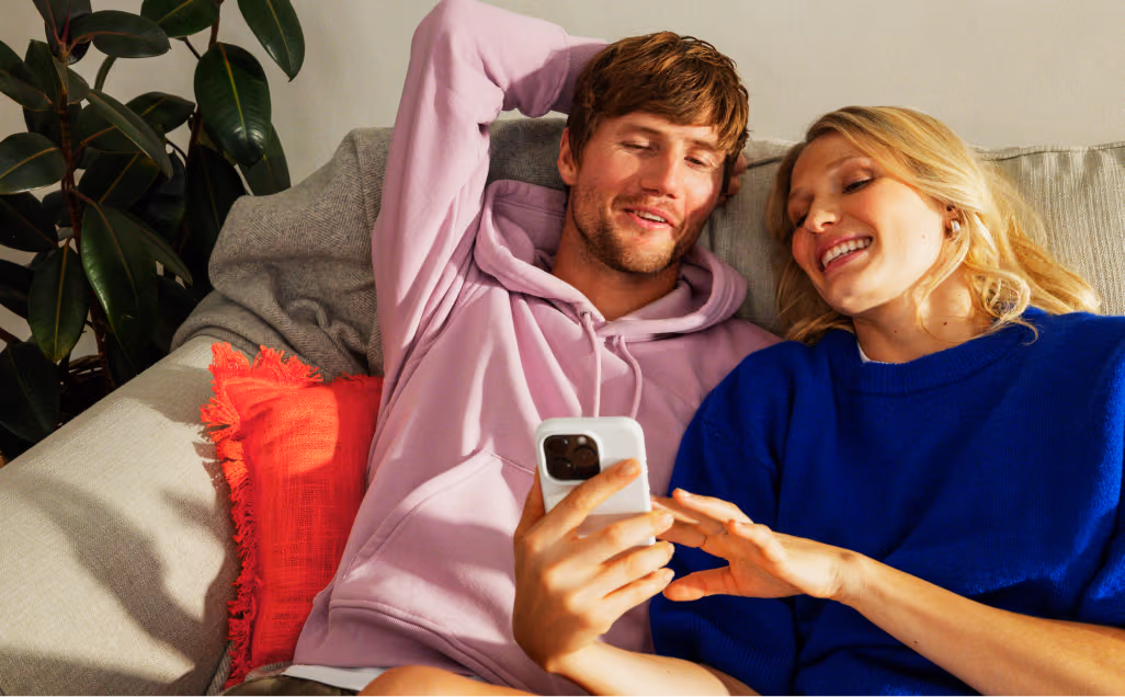 Two people sitting close together on an outdoor lounge sofa, smiling and laughing while looking at something on a smartphone.