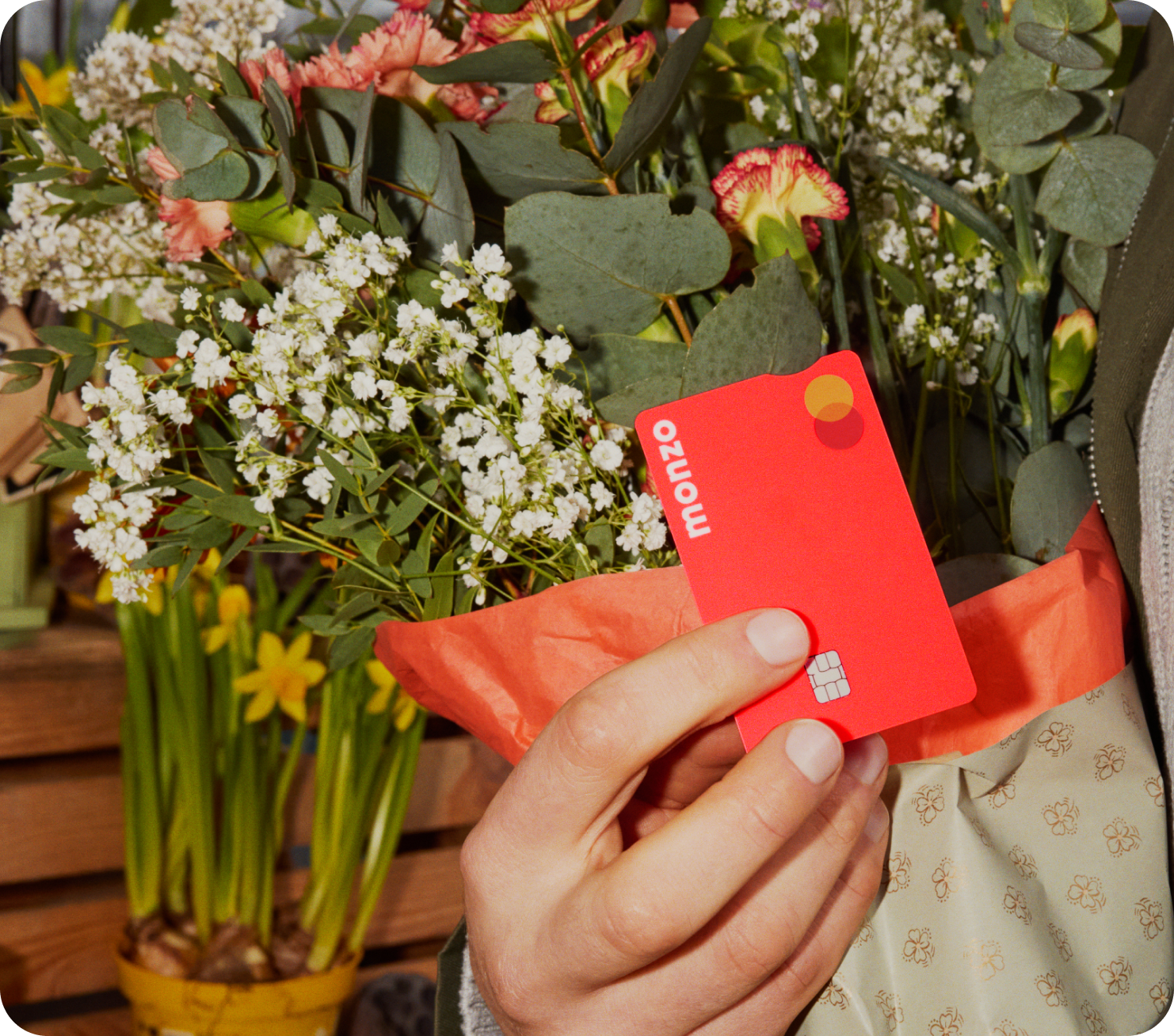 Someone holding their Hot Coral Monzo card against a bunch of bright flowers wrapped in Hot Coral paper.