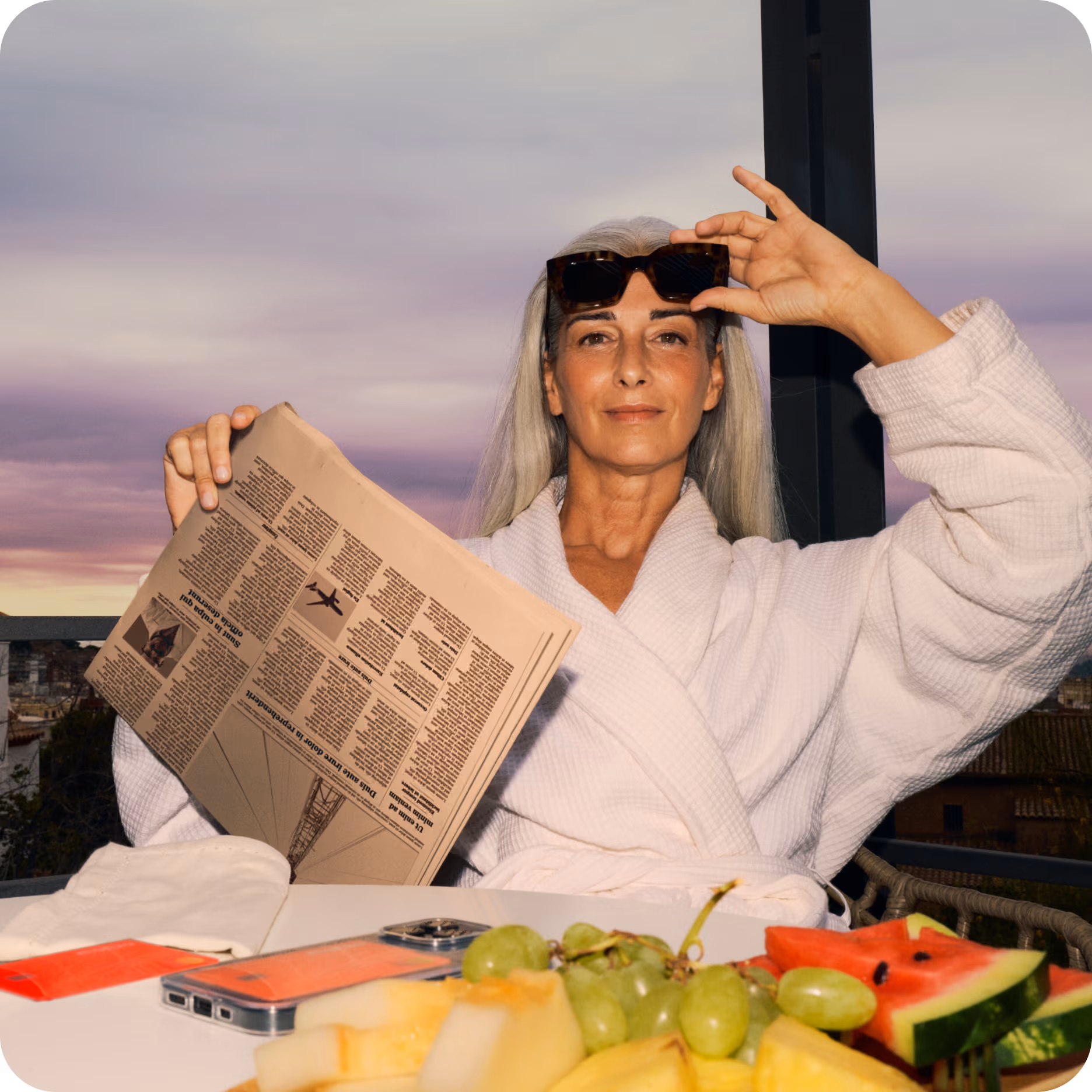Person sitting in front of a fruit platter and reading the newspaper on a balcony wearing sunglasses a robe.