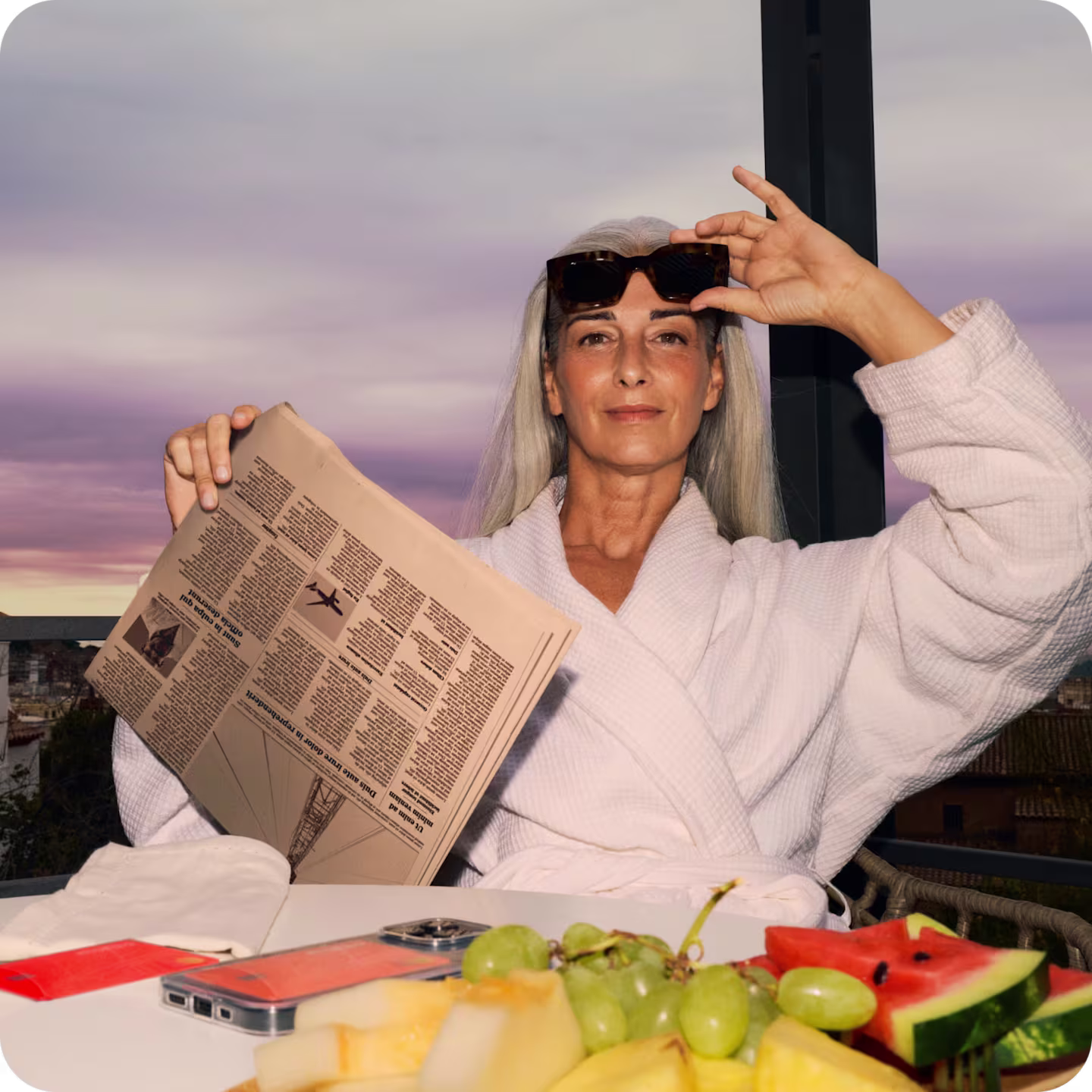 Person sitting in front of a fruit platter and reading the newspaper on a balcony wearing sunglasses a robe.
