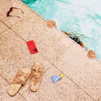 Someone submerges themselves in a swimming pool. On the tiles at the edge of the swimming pool are a pair of sunglasses, a mobile phone with a hot coral phone case, a pair of sandals and a tube of suncream.
