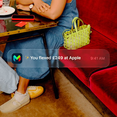 A woman in a blue dress sitting at a table on a red sofa with a green handbag beside her. A phone notification is laid over the image, saying 'You flexed £249 at Apple'