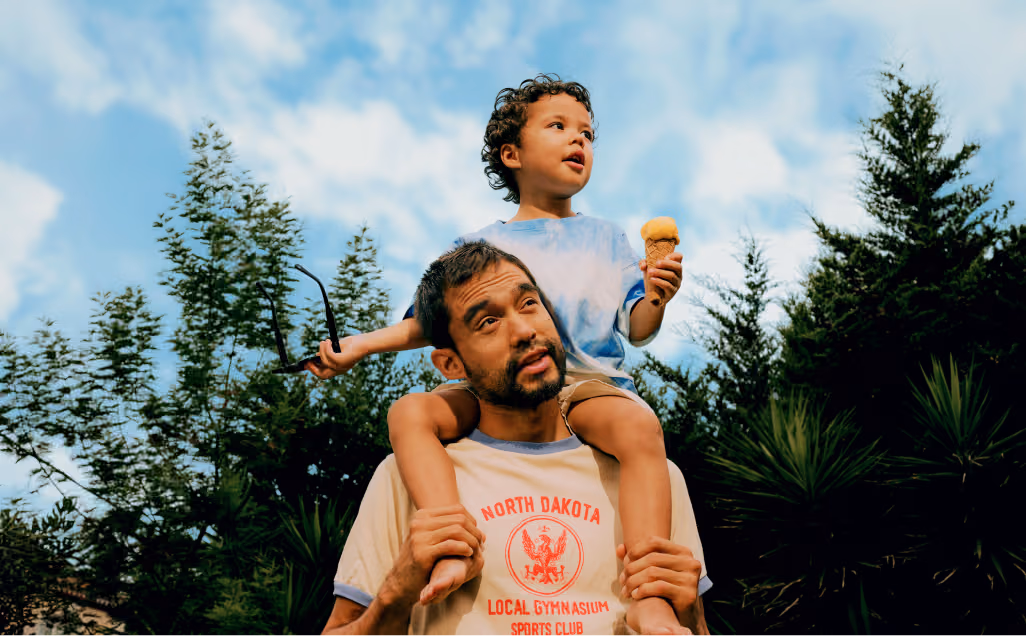 A parent walking through a wooded area with a young child riding playfully on their shoulders.