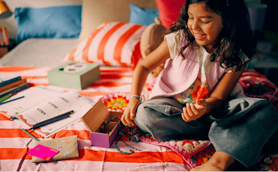 A high-angle shot of a child with dark wavy hair sitting on a colourful striped bed, smiling while playing with a wooden jewellery box. Spread around them