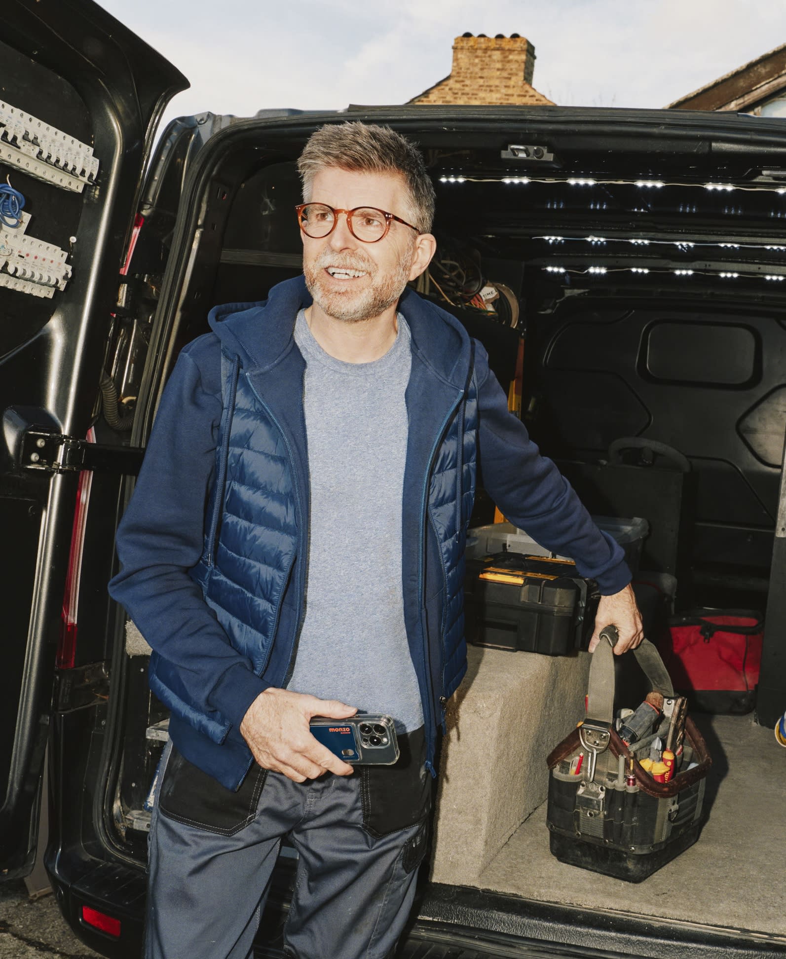 A construction worker in a blue puffer jacket and glasses standing by an open van boot.