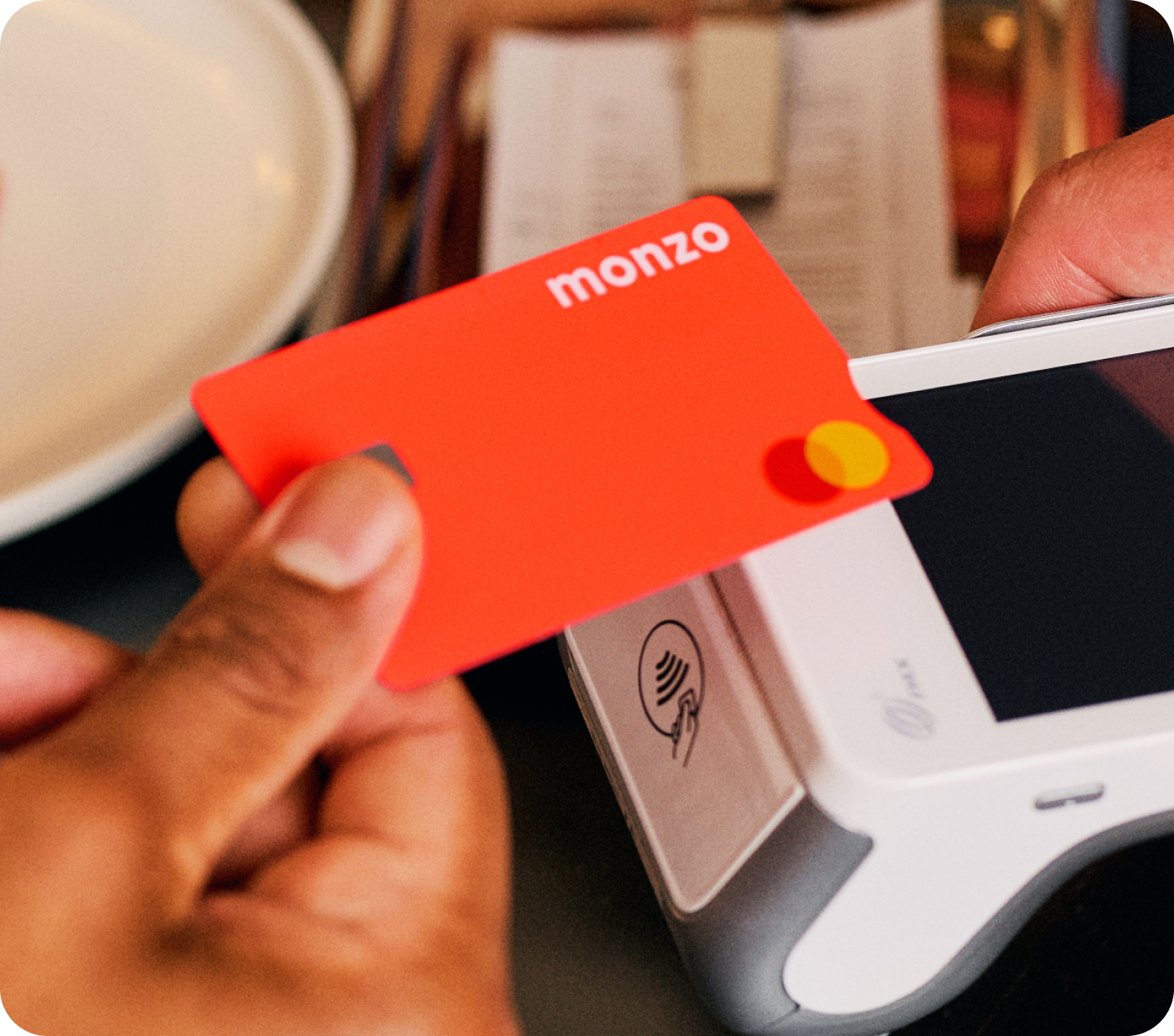 A hand is holding a Monzo Hot Coral card up to a contactless payment terminal.
