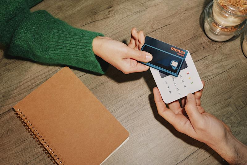 Person in a green jumper tapping a Monzo Expense card on a white card reader at a wooden table, with a notebook and drink nearby.