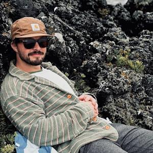 Photo of Steven Jakubowski – he's sat down on a chair wearing a beige cap, black sunglasses and a green and white stripy shirt. 