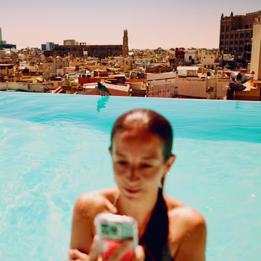 A person in a turquoise infinity pool looks at a smartphone featuring a hot coral card in its case. The background shows a panoramic rooftop view of a sun-drenched city with classical architecture and birds perched on the pool's edge.