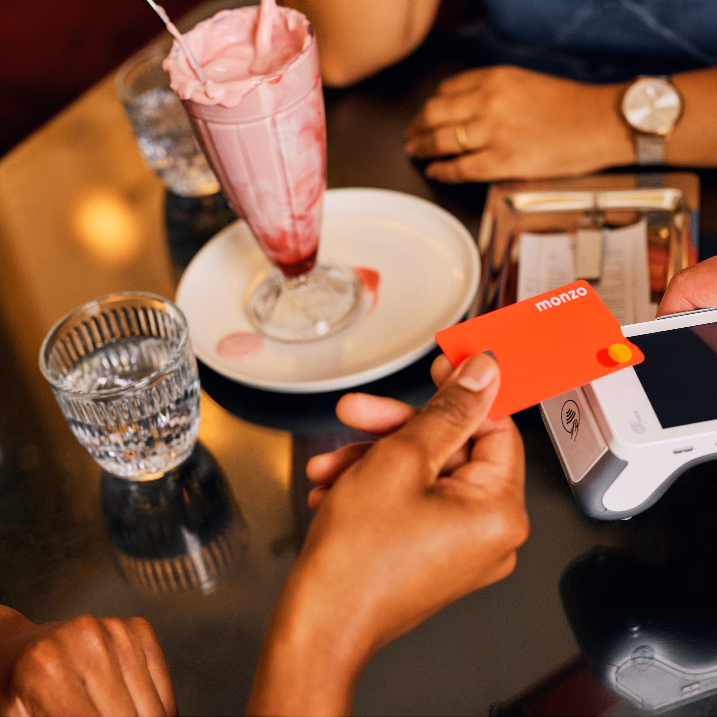 A hand holds a Monzo debit card over a white card reader on a dark table. Nearby, a pink milkshake in a tall glass sits on a white plate, with a ribbed glass of water beside it. In the background, another hand wearing a watch rests on the table.