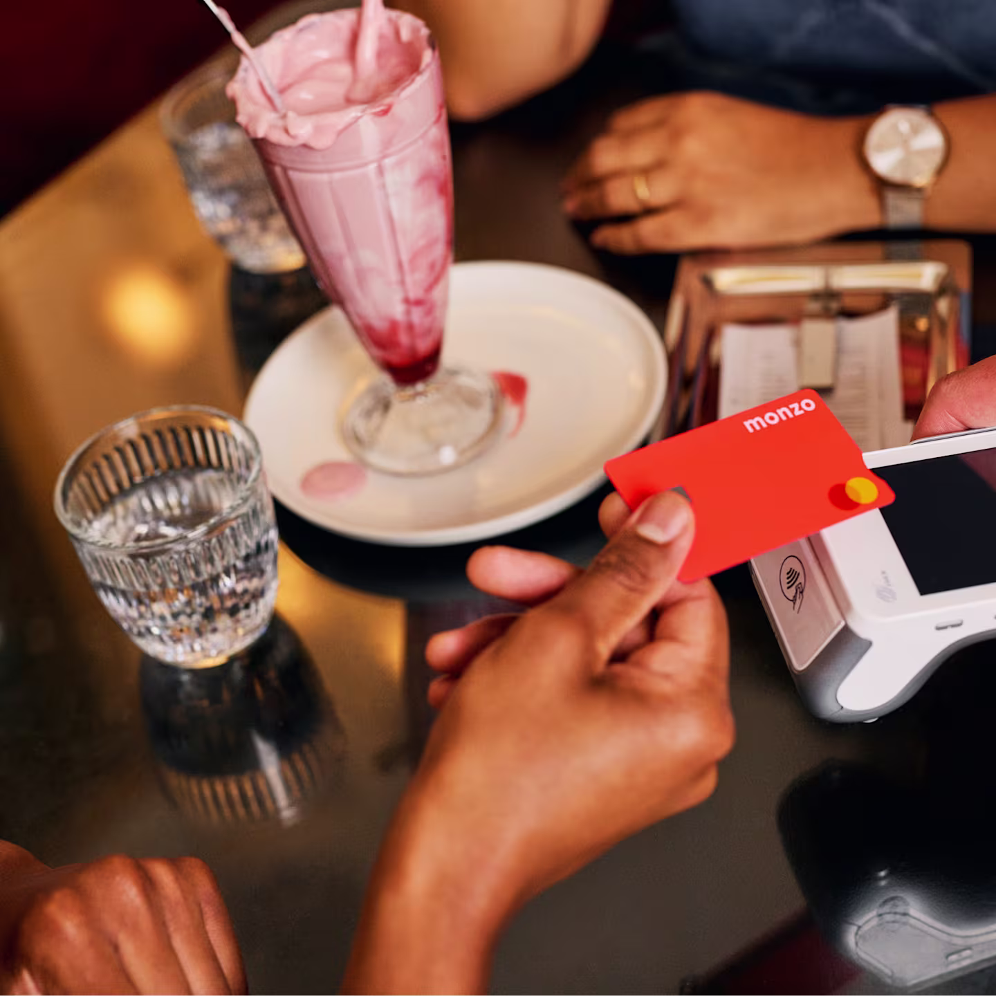A hand holds a Monzo debit card over a white card reader on a dark table. Nearby, a pink milkshake in a tall glass sits on a white plate, with a ribbed glass of water beside it. In the background, another hand wearing a watch rests on the table.