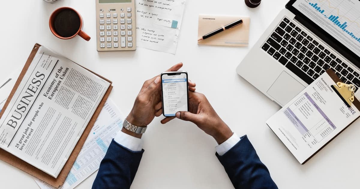 Some hands holding a phone over a desk covered with a newspaper, laptop, calculator, and other papers