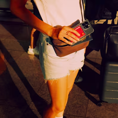 A person standing in a sunlit terminal holds a smartphone with a hot coral Monzo card tucked into the back of the case, held alongside a passport over a brown crossbody bag.