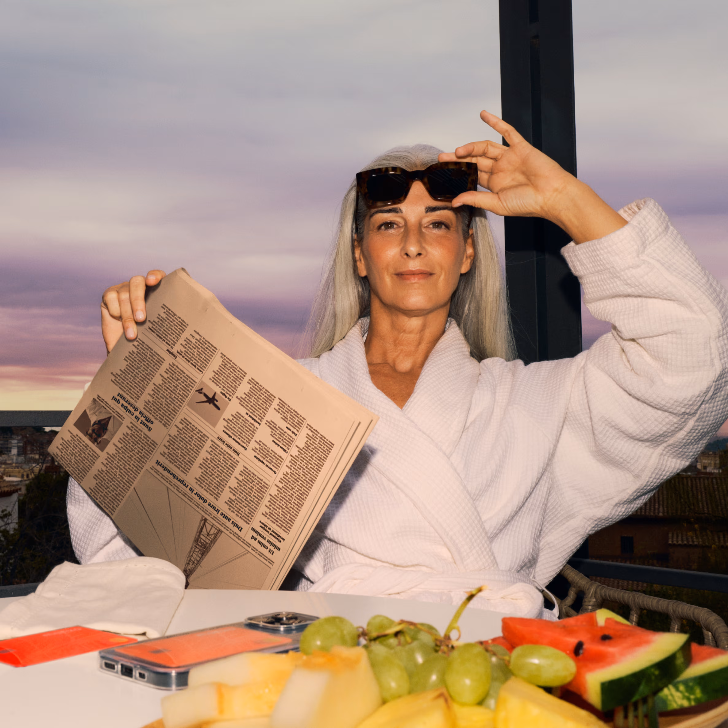A woman sat at a table wearing a white robe and sunglasses, holding a newspaper in her hand. The sun rises in the background and a selection of fruits are arranged on the table in front of her, alongside a mobile phone with a Monzo debit card in the phone case.