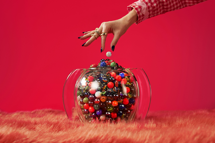 A hand drops a white marble into a glass bowl filled with colorful marbles, set on a pink furry surface.