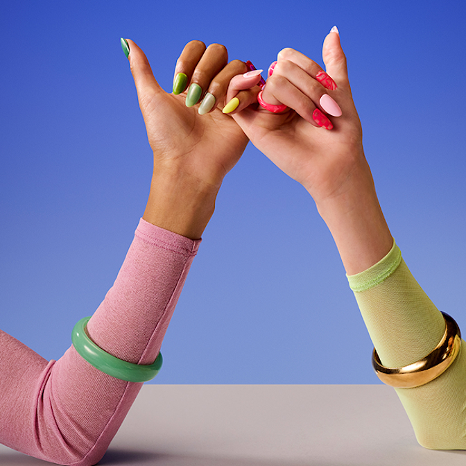 A close-up of two people making a pinky promise against a solid blue and white background. The person on the left wears a pink long-sleeved shirt, a thick green bangle, and has their nails painted in various shades of green. The person on the right wears a pale lime-green long-sleeved shirt, a thick gold bangle, and has their nails painted in shades of pink, red, and yellow.