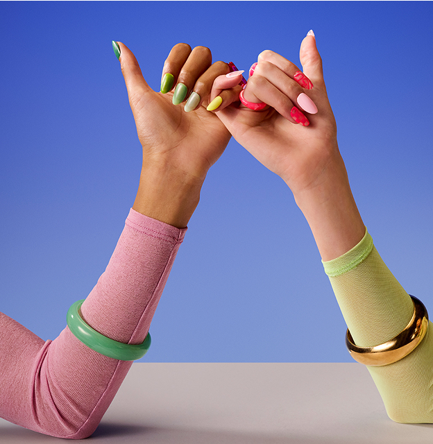 A close-up of two people making a pinky promise against a solid blue and white background. The person on the left wears a pink long-sleeved shirt, a thick green bangle, and has their nails painted in various shades of green. The person on the right wears a pale lime-green long-sleeved shirt, a thick gold bangle, and has their nails painted in shades of pink, red, and yellow.