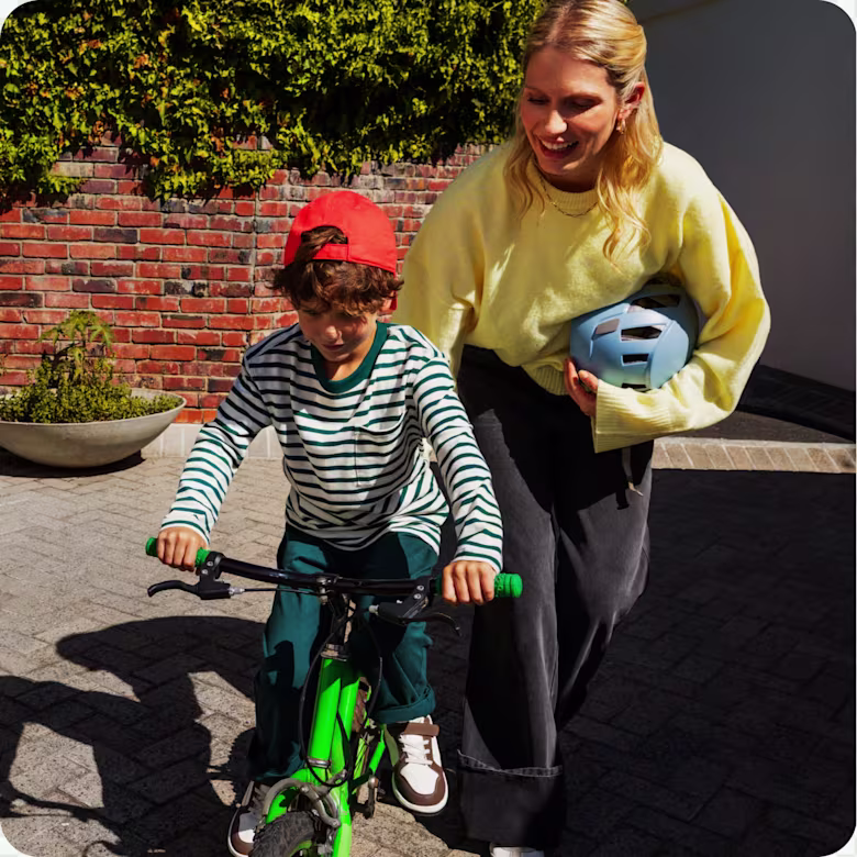 A woman supports a young boy wearing a red cap as he smiles and learns to ride a green bicycle on a brick walkway.