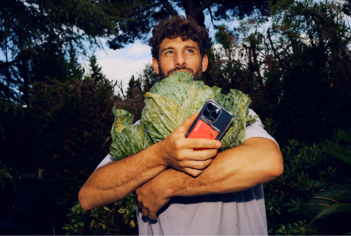 A man stood in an allotment holding in his arms two large, leafy cabbages he has grown. He also holds a mobile phone with a Monzo debit card in the phone case in his hand.