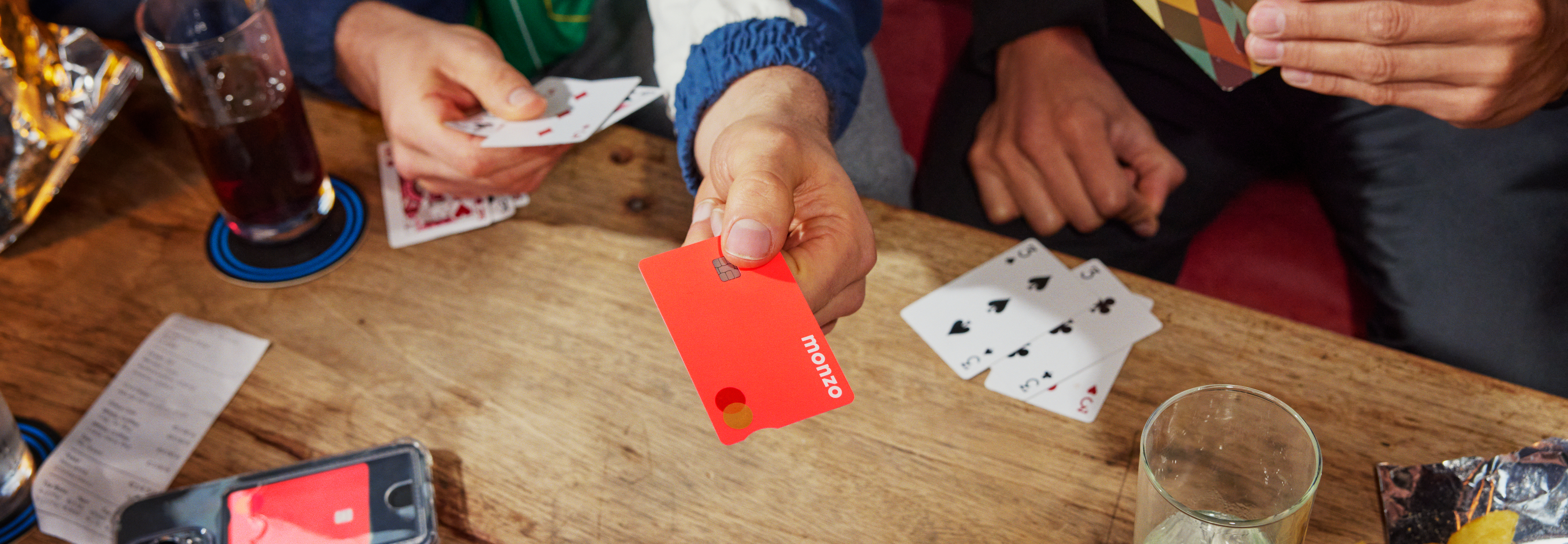 Friends playing cards around a table with drinks, one person holding their coral Monzo card during the game.