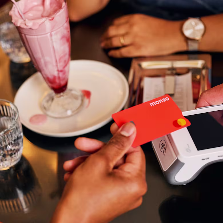 A person taps a hot coral Monzo card on a card reader to make a contactless payment at a café. A pink milkshake is visible on the table in the background.