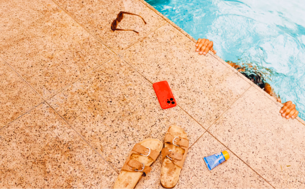 Poolside scene with sandals, a phone, suncream and a person swimming underneath the water.
