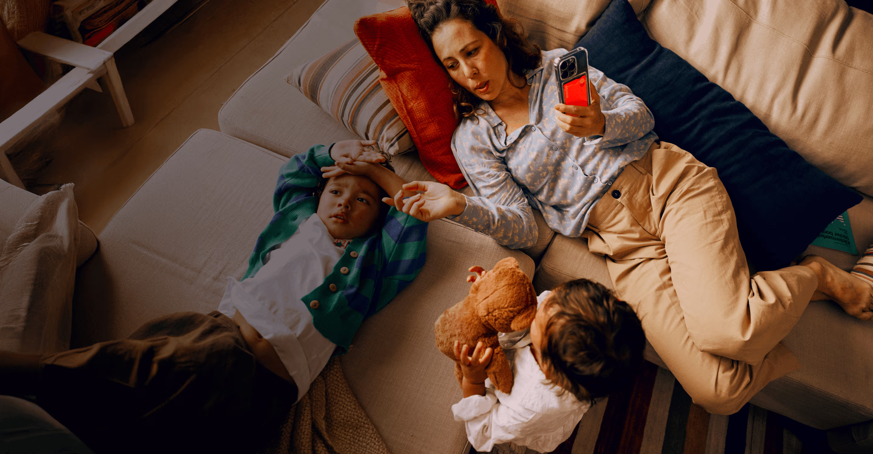 A woman is relaxing on the sofa with her family. She's holding up her phone – it has a clear case on it that holds her Hot Coral Monzo bank card.