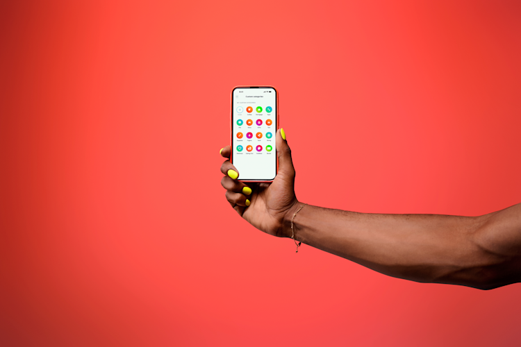A person's arm holds up a smartphone displaying colorful app icons against a coral background.