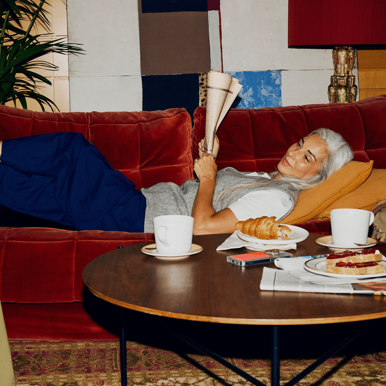 Person with long grey hair reclining on a red velvet sofa, holding a newspaper while breakfast items and coffee sit on a round wooden coffee table in the foreground.