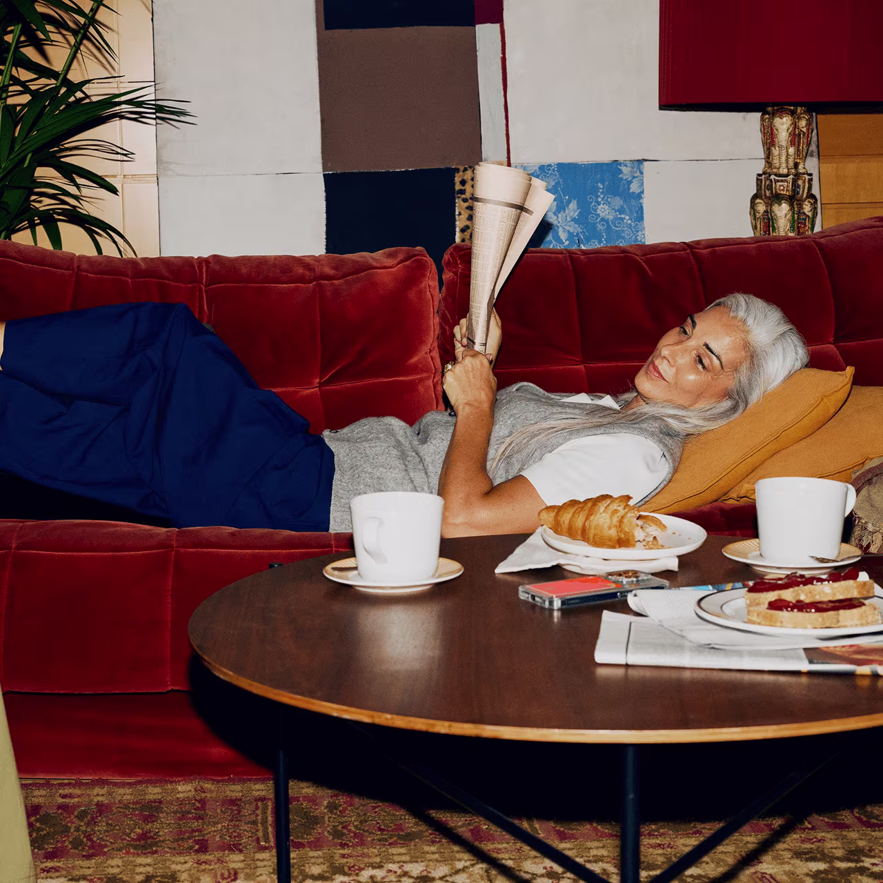 Person with long grey hair reclining on a red velvet sofa, holding a newspaper while breakfast items and coffee sit on a round wooden coffee table in the foreground.