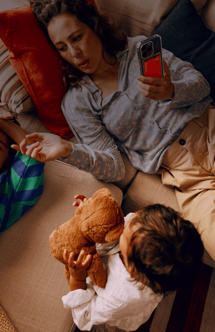 A woman is relaxing on the sofa with her family. She's holding up her phone – it has a clear case on it that holds her Hot Coral Monzo bank card.