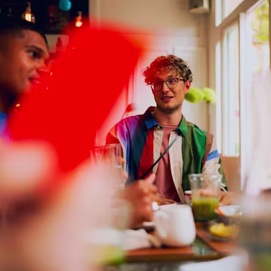 A group of friends sits at a brightly lit cafe table. In the blurred foreground, a hand holds up a hot coral card, while in the background, a person with glasses and a colourful striped shirt smiles during the conversation.