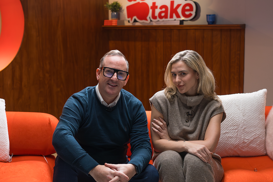 A man and a woman sitting on a hot-coral sofa, looking to camera. 