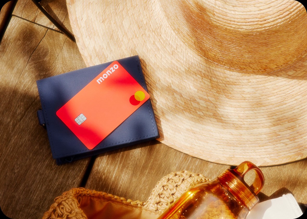 A hot coral Monzo card laid on top of a sun hat, next to a beach bag.