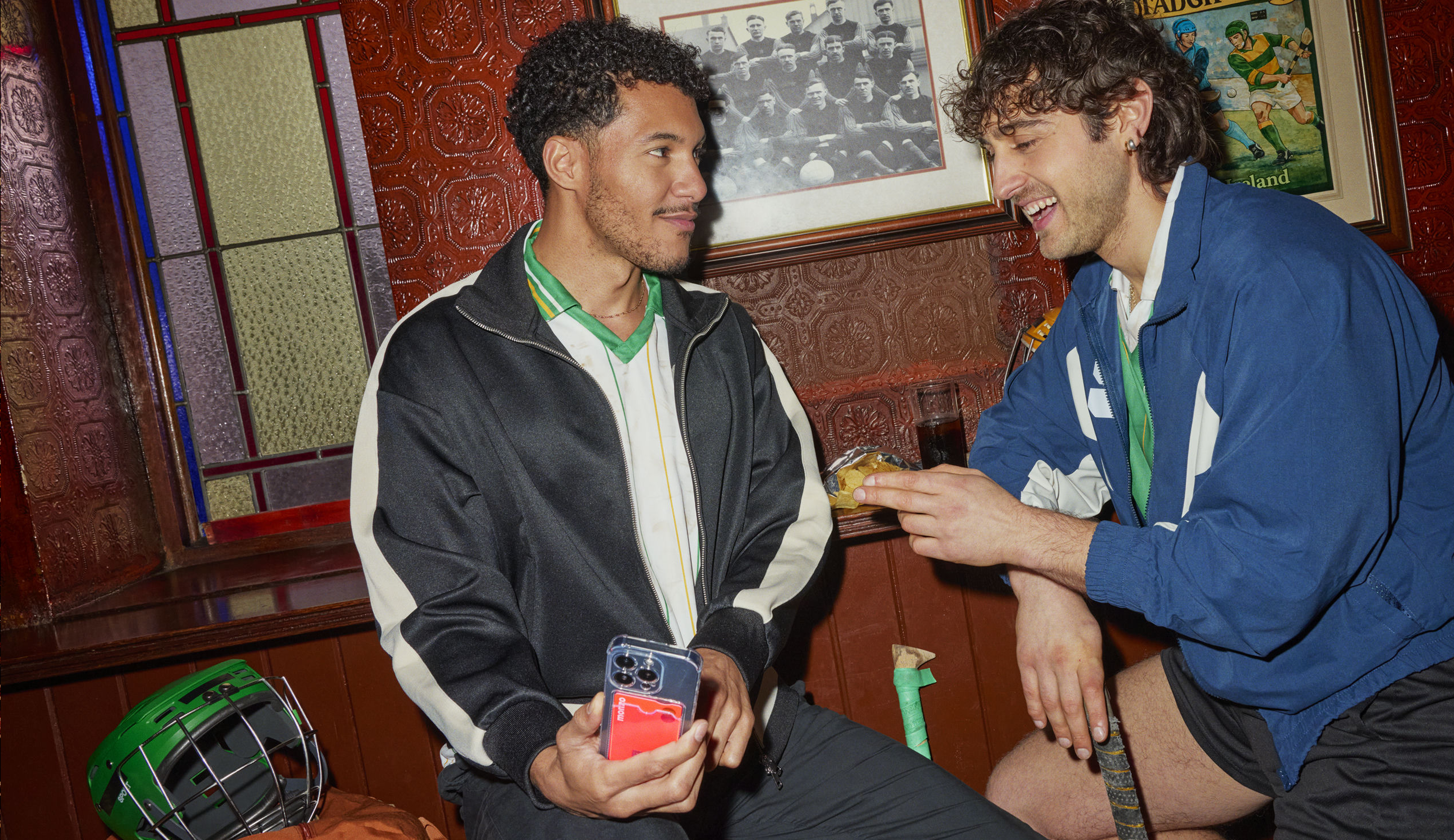Two friends chatting in a traditional pub with vintage football memorabilia on the walls. One holds a phone while the other holds a drink, sharing a lighthearted moment.