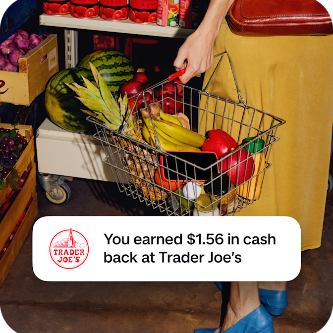 A shopper holds a wire basket filled with fresh pineapple and bananas, overlaid with a notification confirming $1.56 in cash back earned at Trader Joe's.