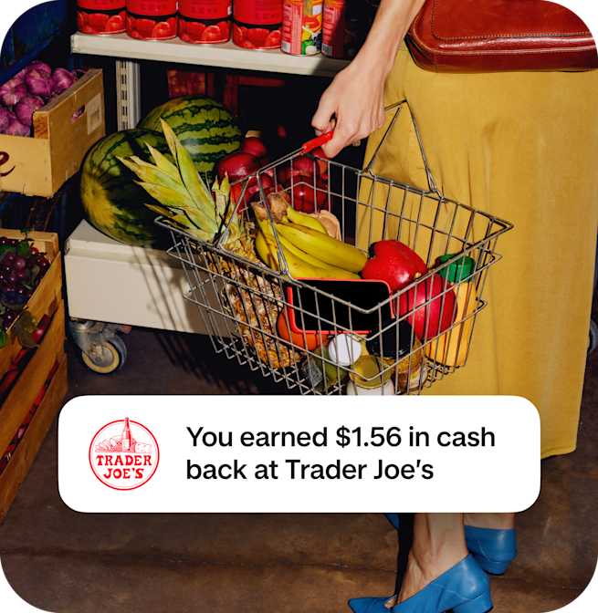 A shopper holds a wire basket filled with fresh pineapple and bananas, overlaid with a notification confirming $1.56 in cash back earned at Trader Joe's.