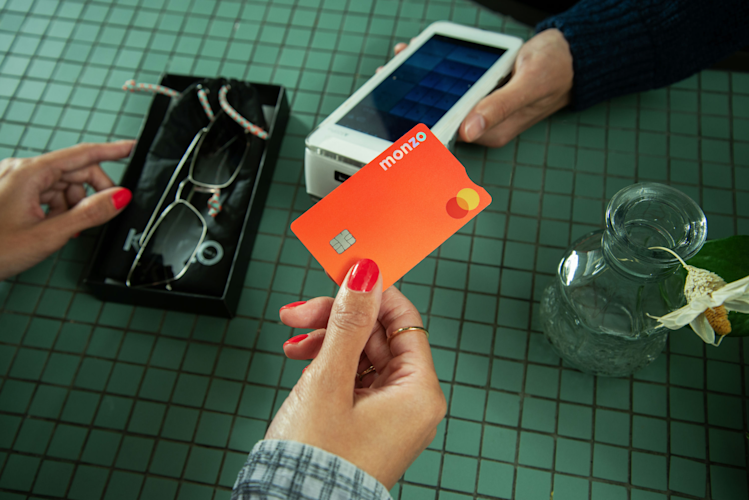 A person's hand paying with an hot coral Monzo card on a payment terminal held by another person.